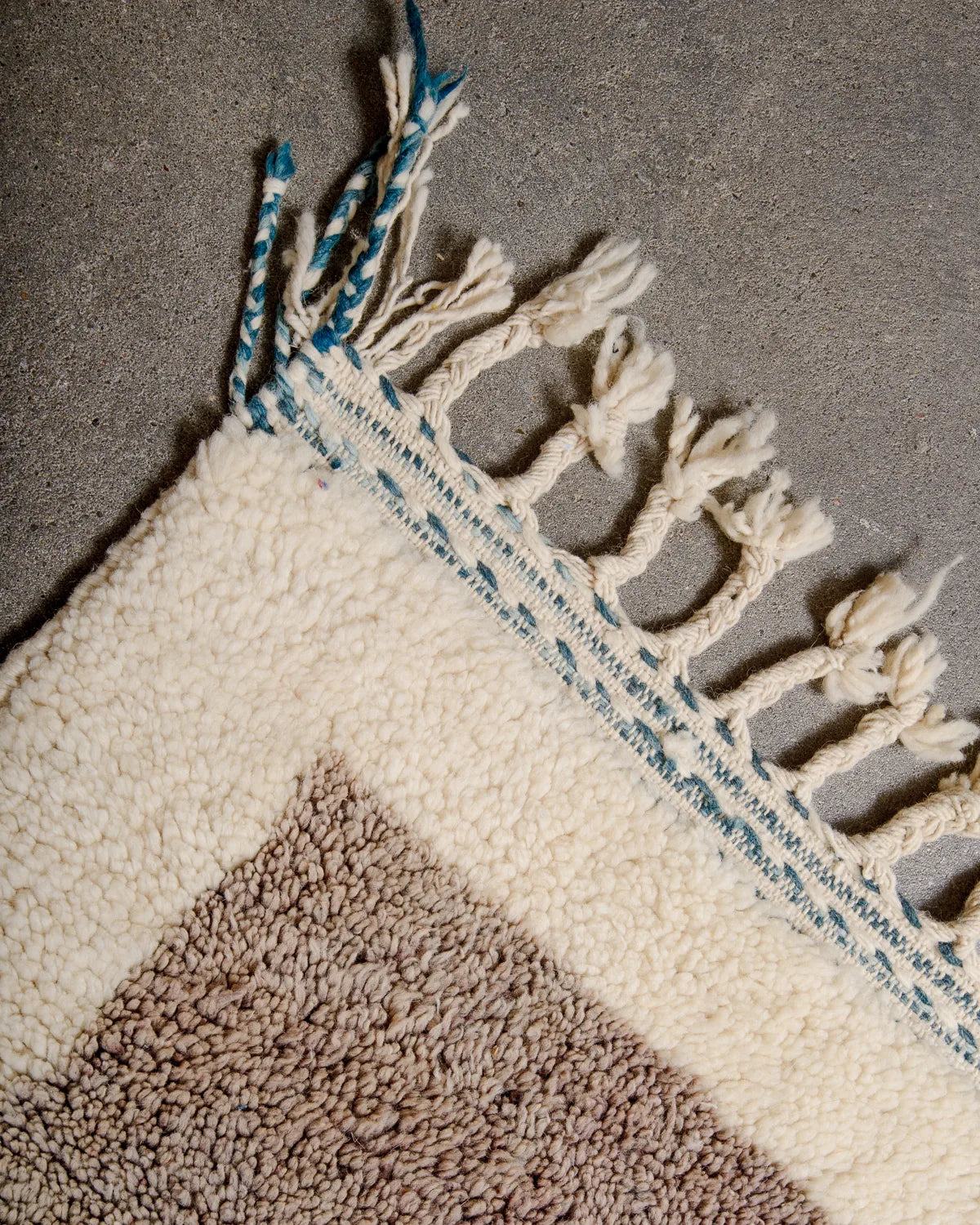 Tassels of edge of wool Berber rug in petrol and natural cream laying on concrete floor.